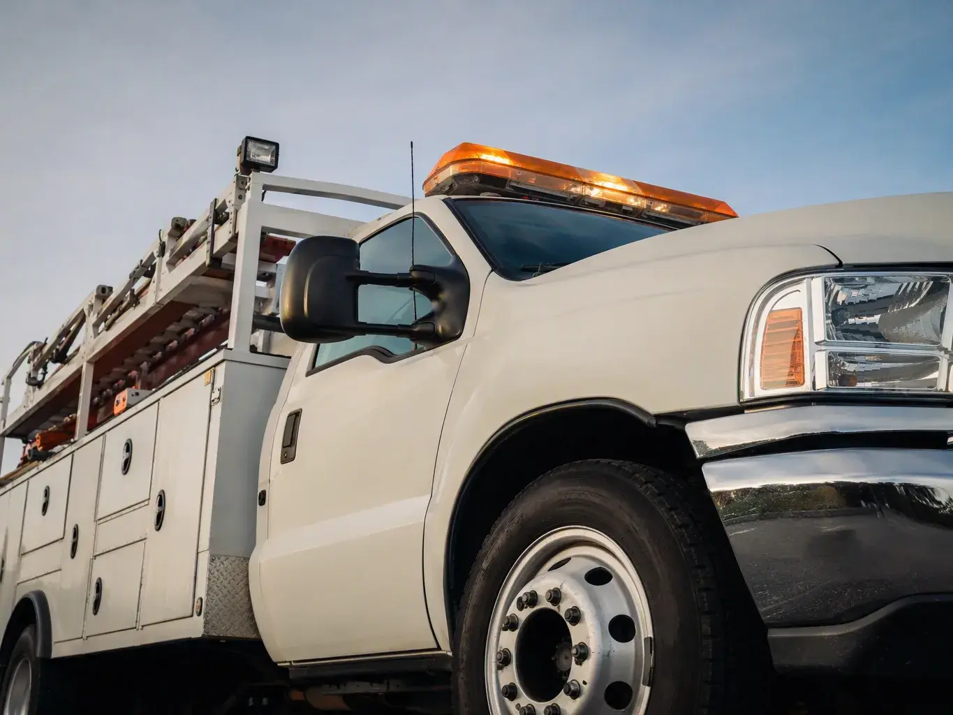 White utility truck with amber light bar and ladder racks parked under a clear