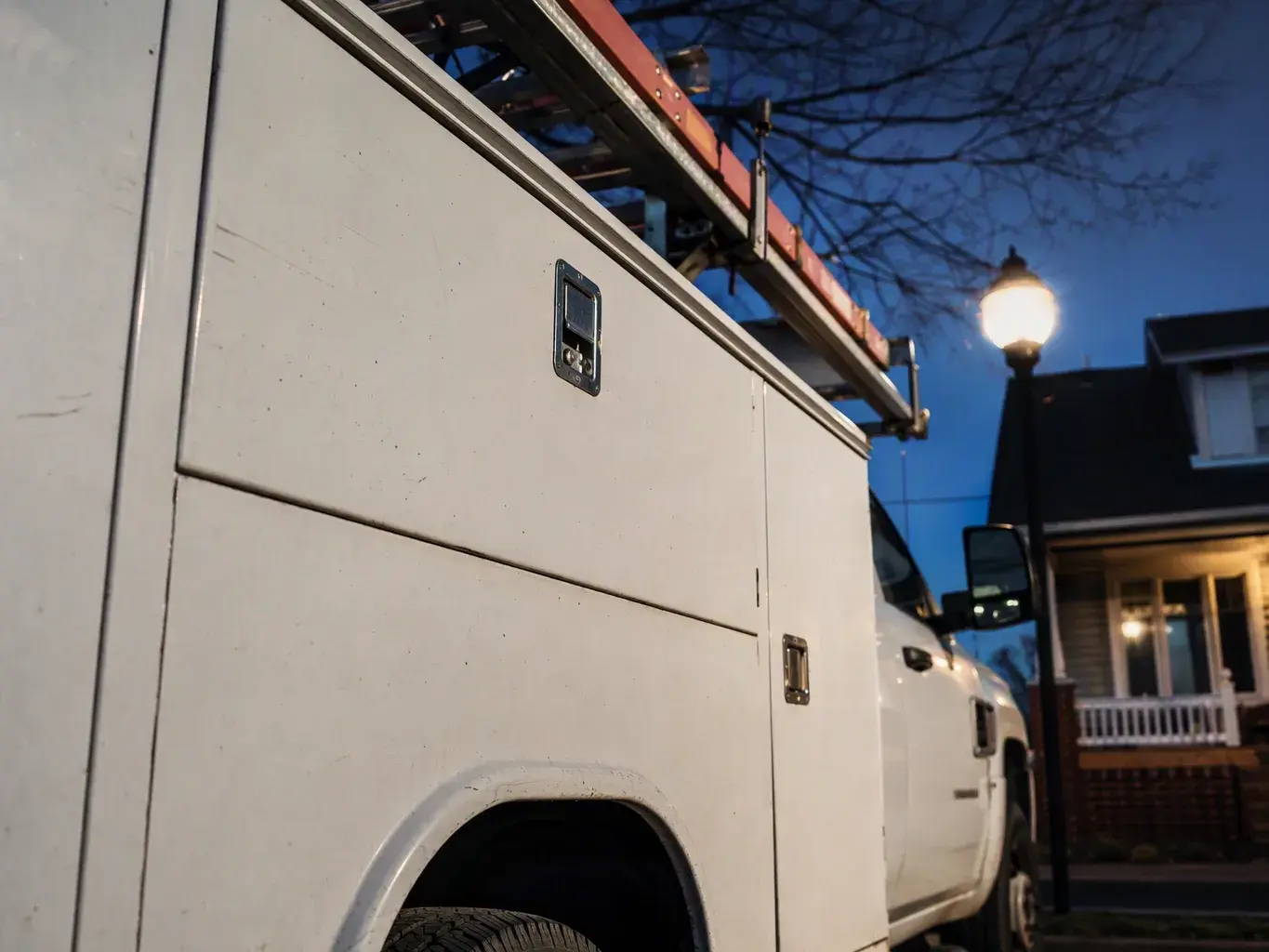 A white utility truck with a ladder rack is parked beside a house at dusk under a lit streetlamp
