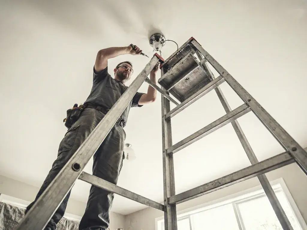 Electrician on a ladder changes a ceiling light in a bright room