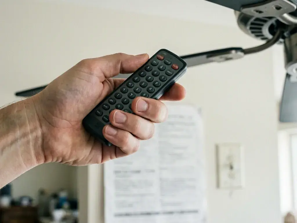 Hand holding a remote control near a ceiling fan in a bright room