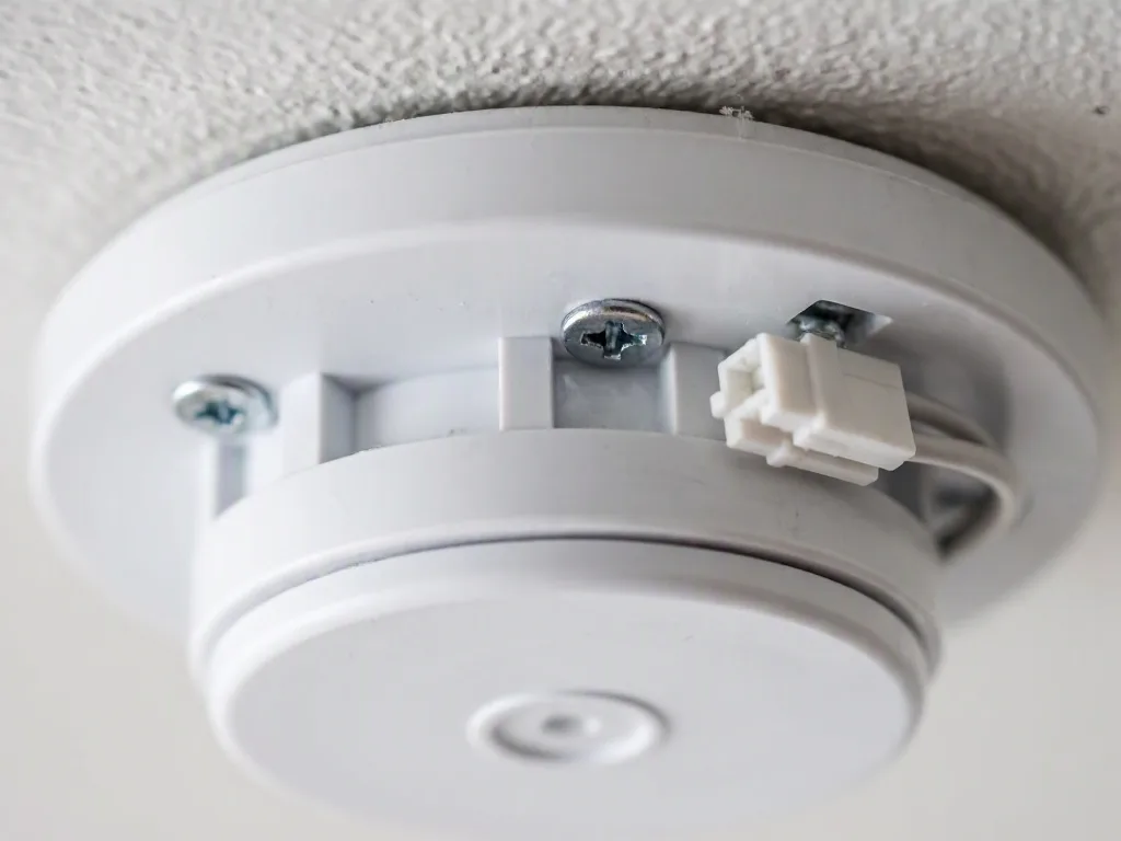 Close-up of a white ceiling-mounted smoke detector with screws and a dangling connector