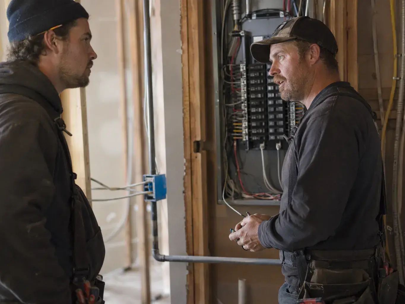 Two workers stand beside an open electrical panel inside a construction wall